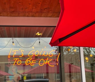 Under a red umbrella, there is a neon orange sign that says “It’s going to be okay!” in a window. In the reflection, there are other red umbrellas and artwork of a woman with blue hair, along with some trees.