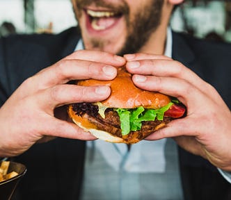 A well-dressed man holds a hamburger, about to chew. He is smiling. The burger is probably not synthetic meat.