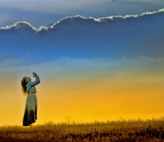 A woman in a field raising her hands in prayer or praise to a sunset-lit sky.