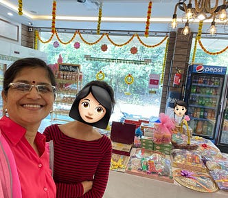 A woman and her daughter, around 15 years old, at a sweet shop. There are hampers of sweets behind them on a large table in the middle of the shop. There are festoons of marigold flowers decorating the shop. A Visicooler with Pepsi in it is at the far end. Another child is in the photo behind the mother and daughter, who is looking at the table with the hampers on it from the far end.