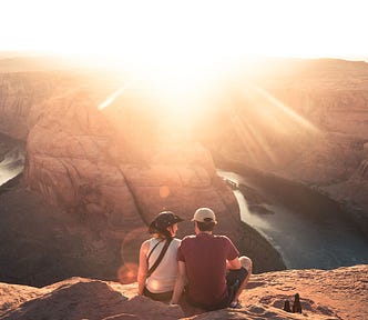 Image of a couple sitting on the rocks looking into the sun and out over a canyon with a river down below.