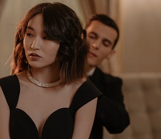 A man in a suit looks intently at an elegantly dressed woman. With her back to him, she leans forward and away and gazes off to the side.
