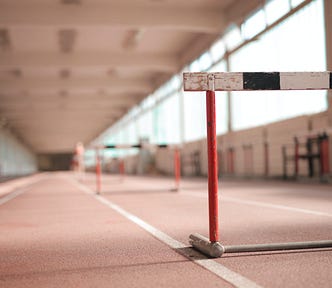 A brown indoor track is lit by daylight. In the foreground, a hurdle sits across one of the track lanes.