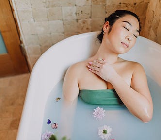 An Asian woman relaxes in a bath with flower tops floating on its surface. Her eyes are closed, and she lovingly holds a washcloth to her chest.