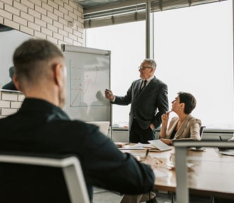A man in a business suit at a whiteboard presenting something, while the rest of the team in business formal attire watches.