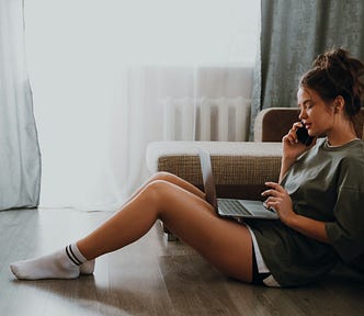 A young woman sits on a wooden floor in front of a sofa. She’s wearing white socks, black shorts and a green sweater over a white t-shirt. She has a laptop on her lap. She is talking to someone on her phone.