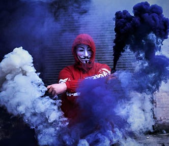 person in red hooded sweatshirt wearing mask and waving blue smoke flares