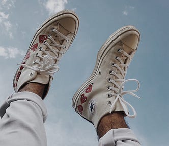 A person’s cuffed jeans and white converse with red heart designs on them raised toward a light blue sky with wispy clouds