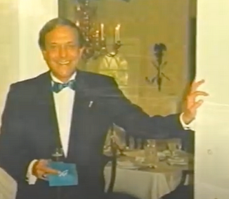 Douglas Marland is wearing a tuxedo with a blue tie, holding a glass of champagne.