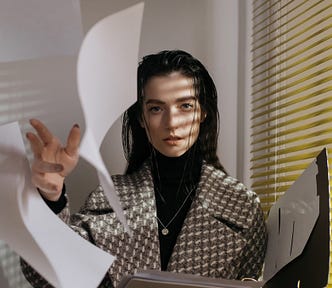 A woman wearing a grey, white, and black checkered coat is looking intently at the camera. She’s throwing papers up in the air and at the camera.