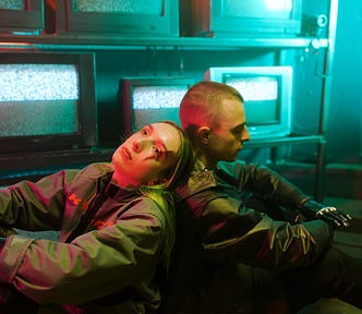 A woman and a man are sitting with their backs to each other in front of wall of TVs displaying static.