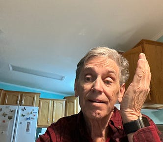 A man with short gray hair wearing a red plaid shirt and a black wristwatch is sitting in a kitchen. He has a slightly puzzled or expressive look, raising one hand beside his face. The background includes wooden cabinets, a refrigerator with magnets, and a light blue wall.