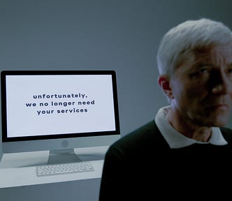 A picture of a man with white hair walking away from his computer. On his screen we read the words “Unfortunately, we no longer need your services”