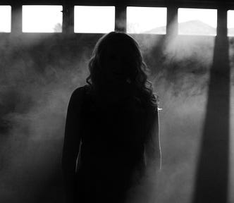 Alt text: A woman stands alone in a dark room, her back to the camera. Light streams through tall windows, casting beams through swirling smoke. Her figure is partially obscured, creating a haunting silhouette.