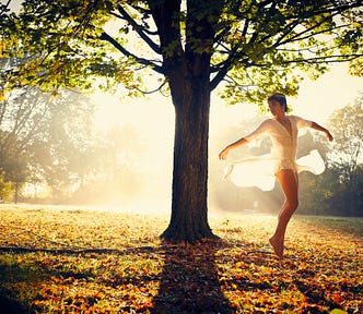 Female dancer dressed in light white clothing, moving in an empty park next to a tree with leaves on an early sunny morning