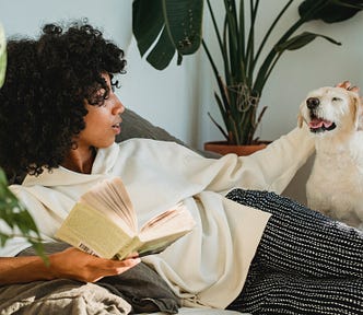 Black woman reclining, holding a book in one hand and petting a white dog with the other hand.