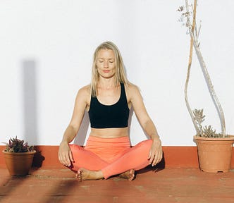 Woman in coral exercise pants sitting with her legs crossed, hands on her knees, and eyes closed.