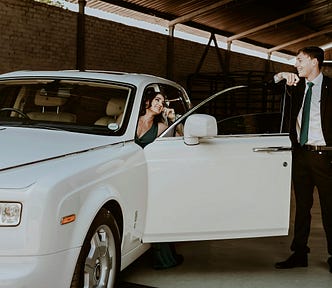 A woman sits inside a rolls-royce, and a man in a suit stands by the car door