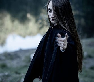 Woman with a gaunt face, long hair, black robes and long black nails reaches out one hand, in the background are trees and a lake