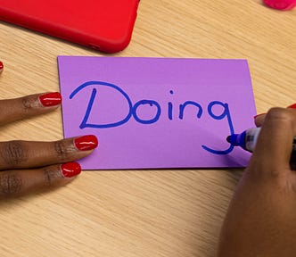 Um post-it roxo com a palavra “Doing” escrita em azul com uma caneta piloto por uma mulher negra com unhas pintadas de vermelho