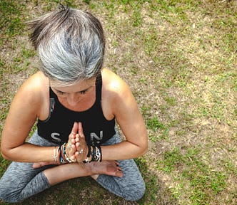 This top view shows a woman with gray hair sitting on the grass, meditating with her hands pressed together in prayer position.