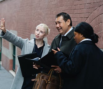 3 people standing on the sidewalk, two women, one male. Both women are talking to the guy in the center, who is holding a folder with some sort of plan. The woman on the left is gesturing towards something to explain something, and the woman on the right is pointing to something in the plan to give emphasis. The man in the center is smiling while looking at what the woman is pointing at.