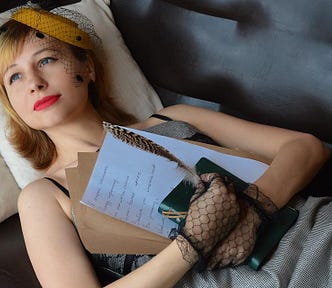 Photo of a young woman lying on a sofa and holding a journal, a feather pen, and a book to her chest as she gazes off into the distance with a dreamy look.