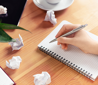 A person with a pen poised to write on blank paper in a notebook with 4 crumpled up sheets of paper next to it.