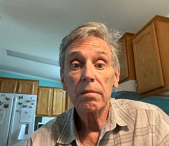 A man with gray hair wearing a plaid shirt looks directly at the camera with a neutral, slightly surprised expression.