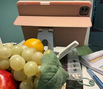 A close-up view of a table with various items, including an iPad in a pink case on a stand, a pile of green grapes, a red apple, an orange, a white cloth napkin with a straw, a white pen, a daily pill organizer, and some papers with highlighted text. The background shows a green wall and part of a doorway.