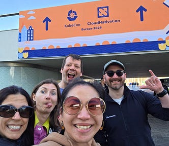 Five people smile for a selfie outdoors in front of a large orange “KubeCon + CloudNativeCon Europe 2026” event banner decorated with stylized buildings, tulips, a bicycle, and a bridge.