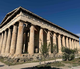 A well‑preserved ancient Greek temple with tall Doric columns stands in an open archaeological site, surrounded by small trees and a gravel path.