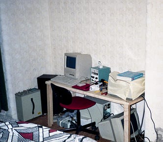 A Mac SE on a homemade table in a bedroom with other equipment nearby such as an oscilloscope, bench power supply, PC, and monitor.