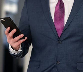 A man wearing a blue suit, holding his phone up as though he is looking at it. The photo is cropped to show only his torso.