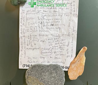 sheet of branded notepad paper held down by a couple of rocks and a shell fragment. A cross and “EMERGENCY AMBULANCE SERVICE” is printed in green across the top of it; writing in black ballpoint pen is all over the page, up and down and sideways.