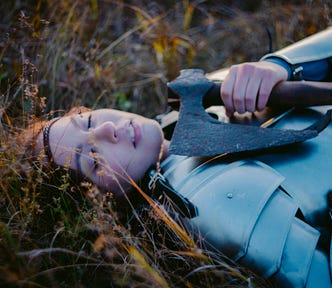 A young woman in full plate armour but no helmet, lying on the ground with her eyes closed.
