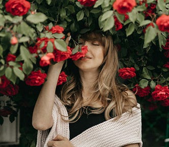 A woman with long brown hair smells rose on a bush just above her.