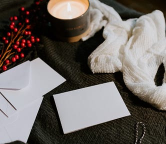 Photo on a dark background of a table, with white envelopes and note cards in the center of the photo. A white scarf lays above the cards, to the right of the photo. A candle in a tin is lit, to provide ambience. A bough of red berries decorates the table.