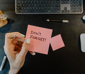A hand holding a post it with “Don’t Forget!” written on it in front of a keyboard and mouse.