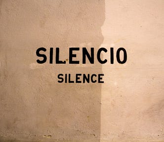 An off-white, roughly plastered wall, with the words ‘silencio’ and ‘silence’ stencilled onto it in black paint.