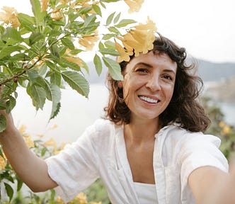 A photo of a woman with dark hair and light brown skin smiling next to a tree with yellow flowers.
