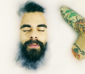 A young, bearded man in a bath of milk. Only his face and part of one arm are visible above the milk. He has his eyes closed.