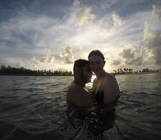 Couple in the ocean, the sunset is happening behind them, the sky is blue, with lots of fluffy clouds.