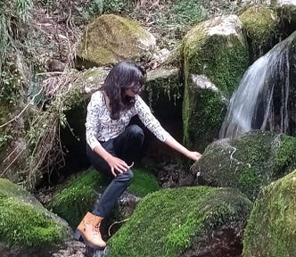 A woman sitting on mossy rocks near a small waterfall, gently touching the water in a quiet forest setting.