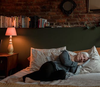 An older woman with grey hair sleeping on top of her bed with all her clothes on in a dimly lit room.