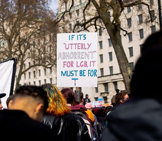 Photograph of a sign at Ban conversion therapy for all — The Trans Rights Protest London, April 2022 that reads “If it’s utterly abhorrent for LGB, it must be for T” in the transgender flag colors.