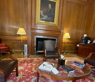 The inside of the Member Lounge at the American Museum of Natural History. Wood-panelled walls with a grand portrait above a fireplace and antique furniture on a beautiful red, floral carpet.