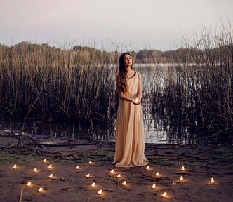 A woman in a white dress stands on the muddy bank of a lake. Lit candles are at her feet. The flora in the background hints the picture was taken in late fall. | Photo courtesy: Carolina Basi, Pexels