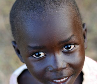 A close up of the face of a young African child in a loose white top, smiling directly at the camera.