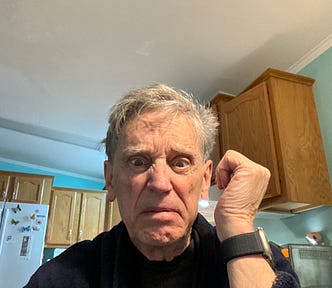 An older person with gray hair looking angry or frightened. The photo is taken in what appears to be a kitchen with wooden cabinets and a refrigerator visible in the background. The kitchen has light blue/mint green walls.